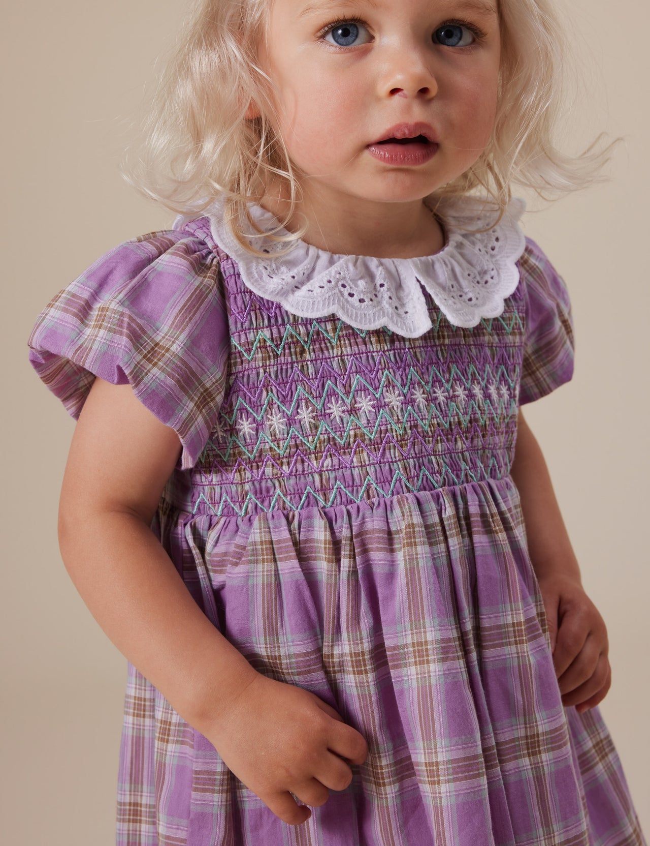 The Goldie + Ace Mauve Check Flo Smocked Dress in Mauve Check pictured on a grey background. The material is cotton. It is a dress made for babies, toddlers and kids.