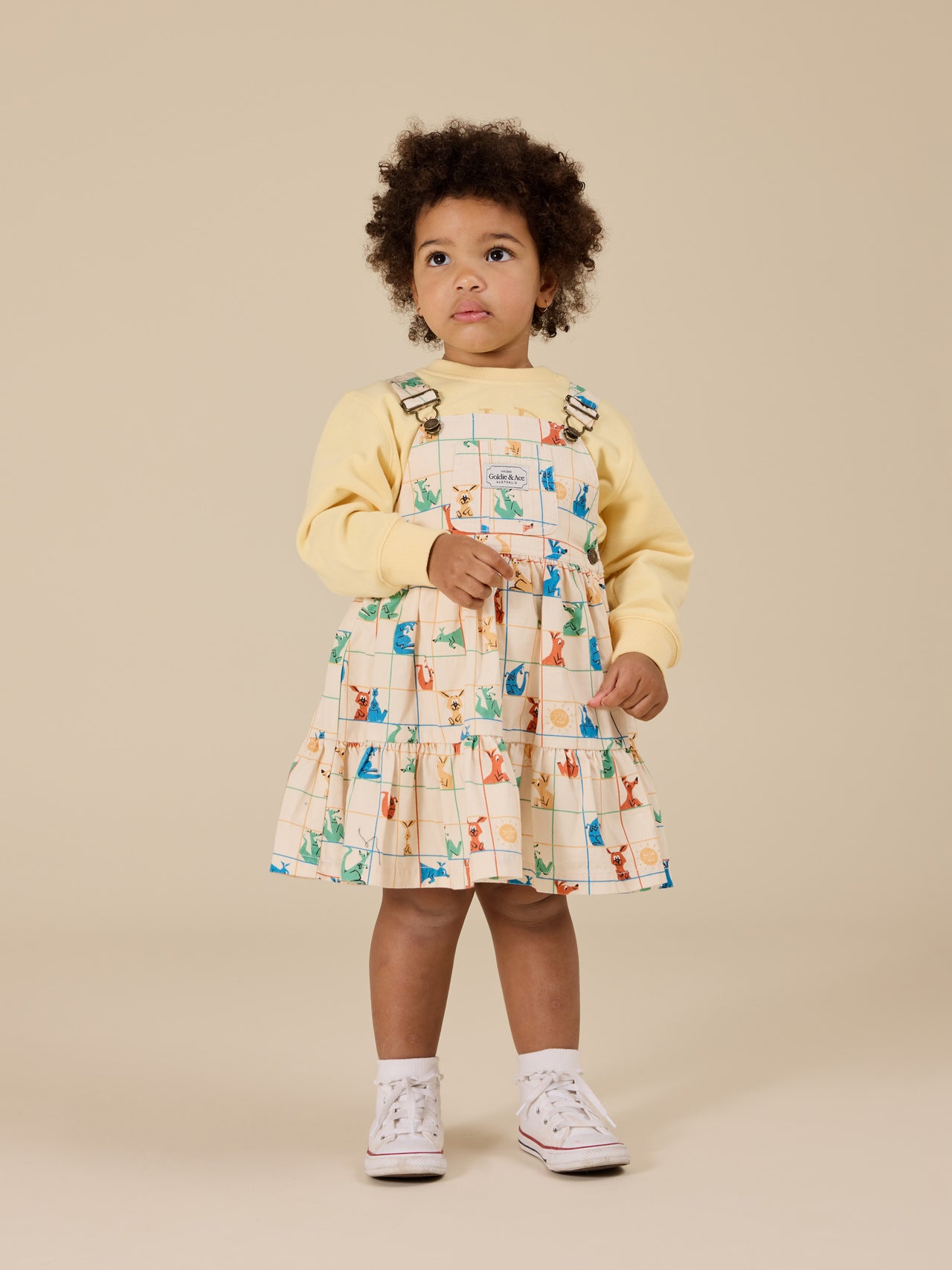 The Goldie + Ace Hopscotch Tully Tiered Pinafore Dress in Vanilla/Primary pictured on a transparent background. The material is 100% cotton. It is a dress made for babies, toddlers and kids.
