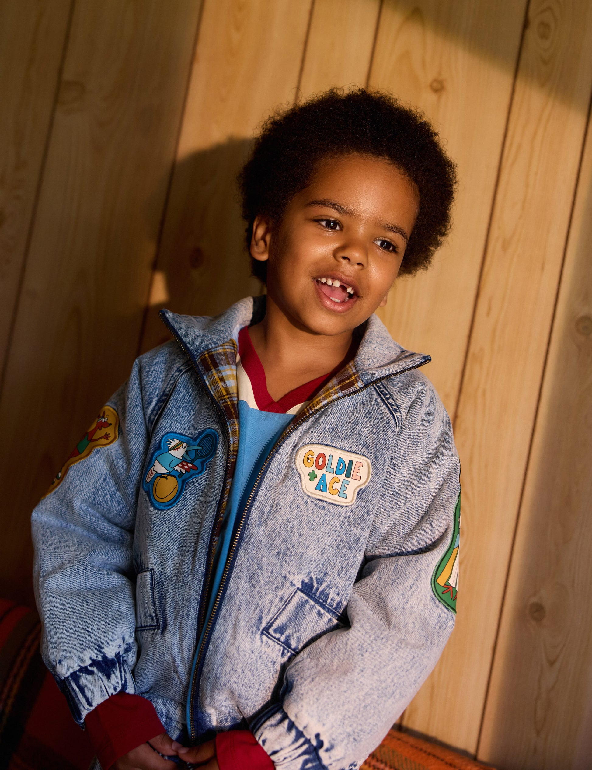 A child wearing a blue denim jacket with patches and text on it, over a red shirt, against a wooden backdrop.