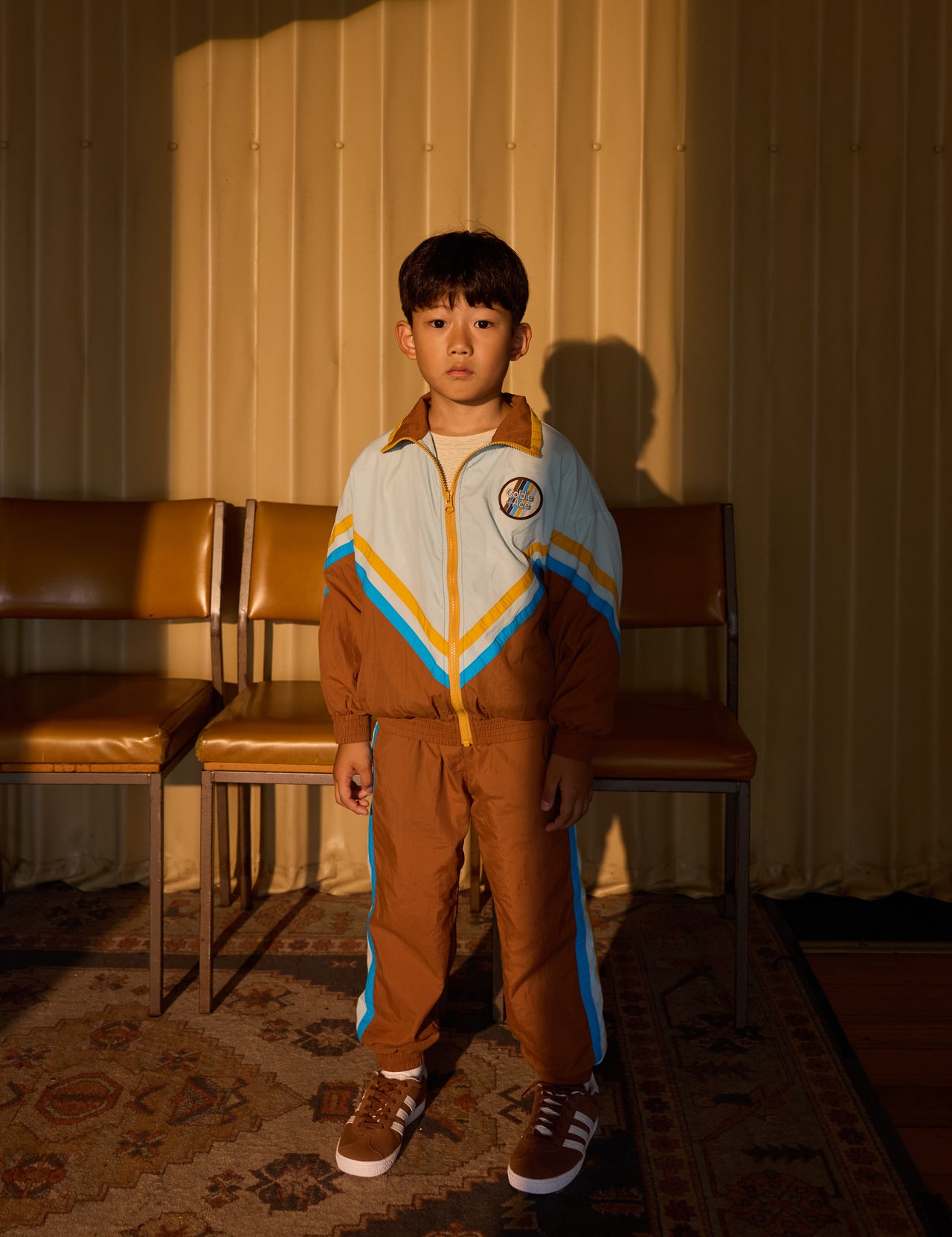 The Goldie + Ace Retro Lightweight Spray Jacket in Brown/Dusty Blue pictured on a grey background. The material is poly with poly lining. It is a jacket made for babies, toddlers and kids.