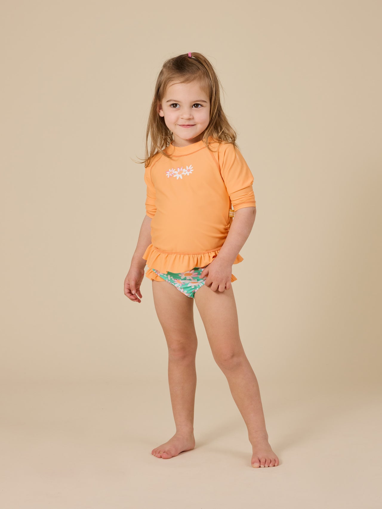 The Goldie + Ace Daisy Chain Frill Rashguard in Orange pictured on a transparent background. The material is . It is a swimwear made for babies, toddlers and kids.