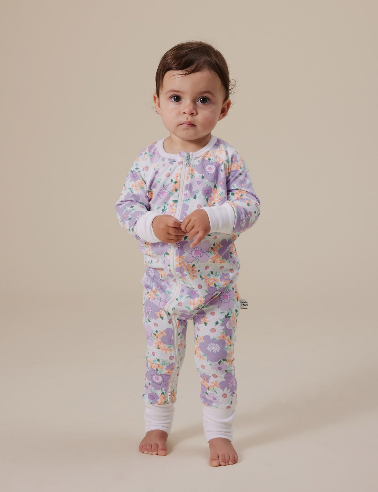 The Goldie + Ace Flora Zipsuit in Lilac Multi pictured on a grey background. The material is soft cotton elastane. It is a babywear made for babies, toddlers and kids.