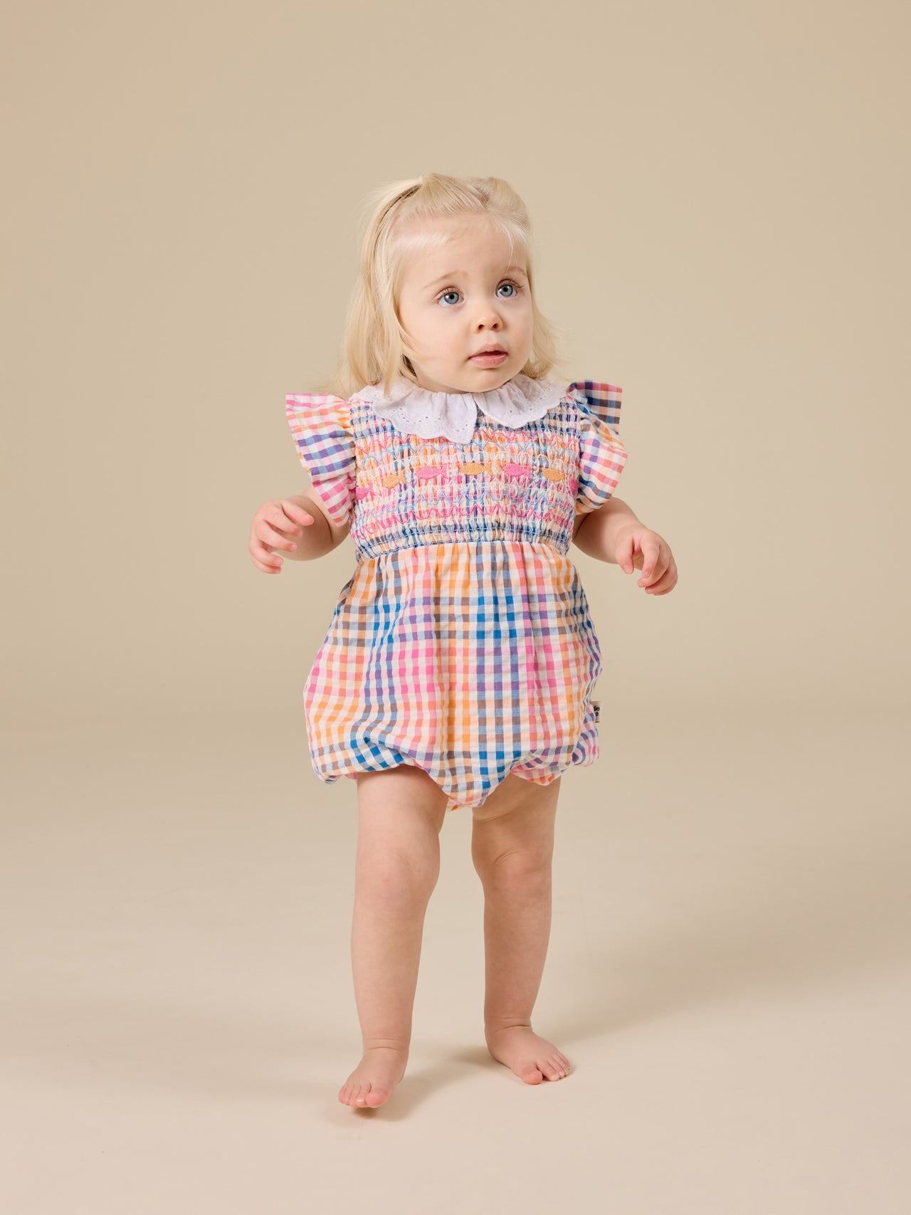 The Goldie + Ace Rainbow Gingham Lani Smocked Romper in Multi pictured on a transparent background. The material is cotton with cotton lining. It is a romper made for babies, toddlers and kids.