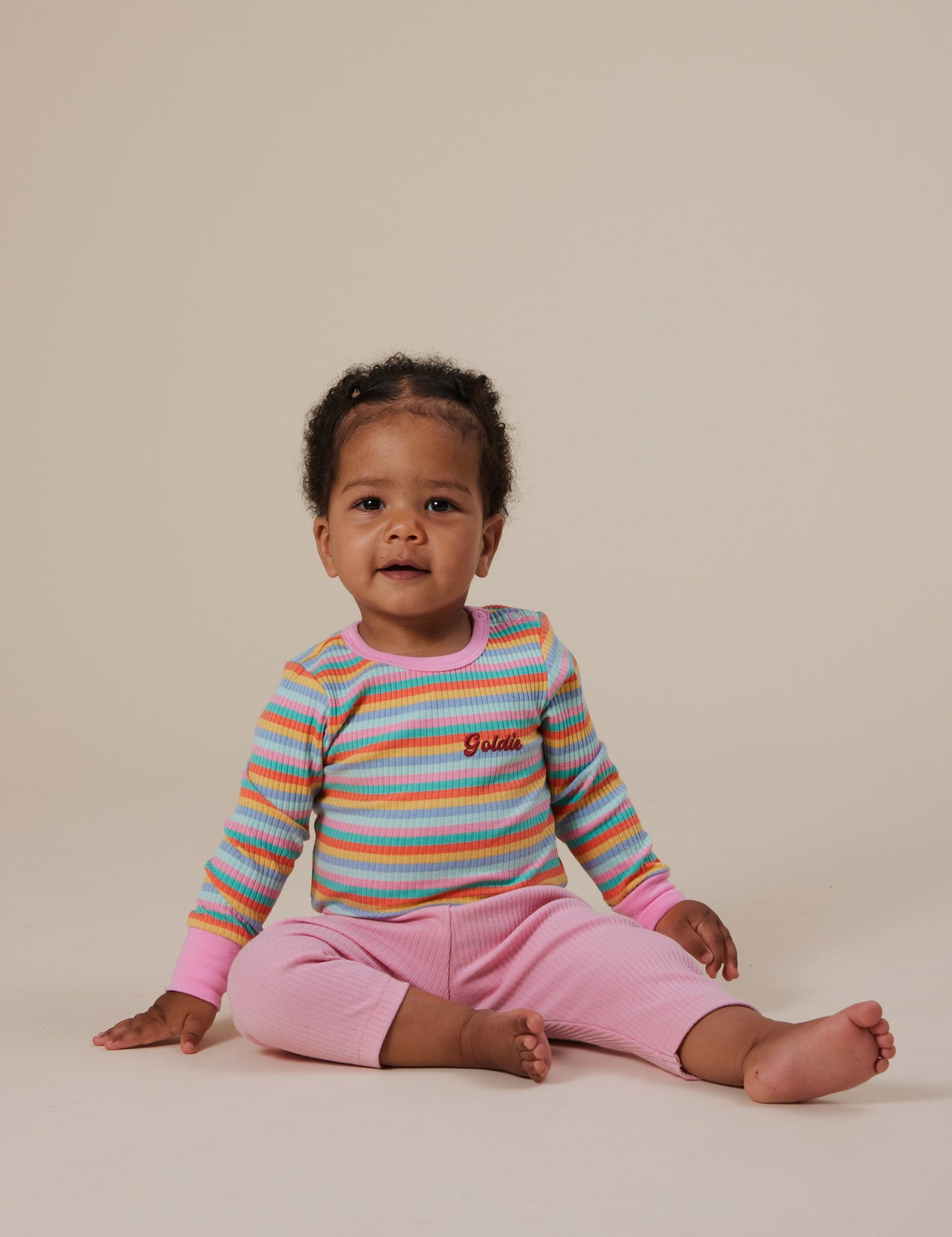 The Goldie + Ace Long Sleeve Rib Bodysuit in Candy Stripe pictured on a grey background. The material is soft ribbed cotton elastane. It is a babywear made for babies, toddlers and kids.