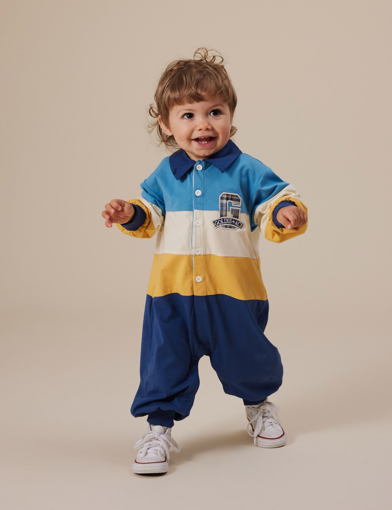 The Goldie + Ace Panelled Rugby Romper in Blue/Golden pictured on a grey background. The material is cotton jersey. It is a romper made for babies, toddlers and kids.