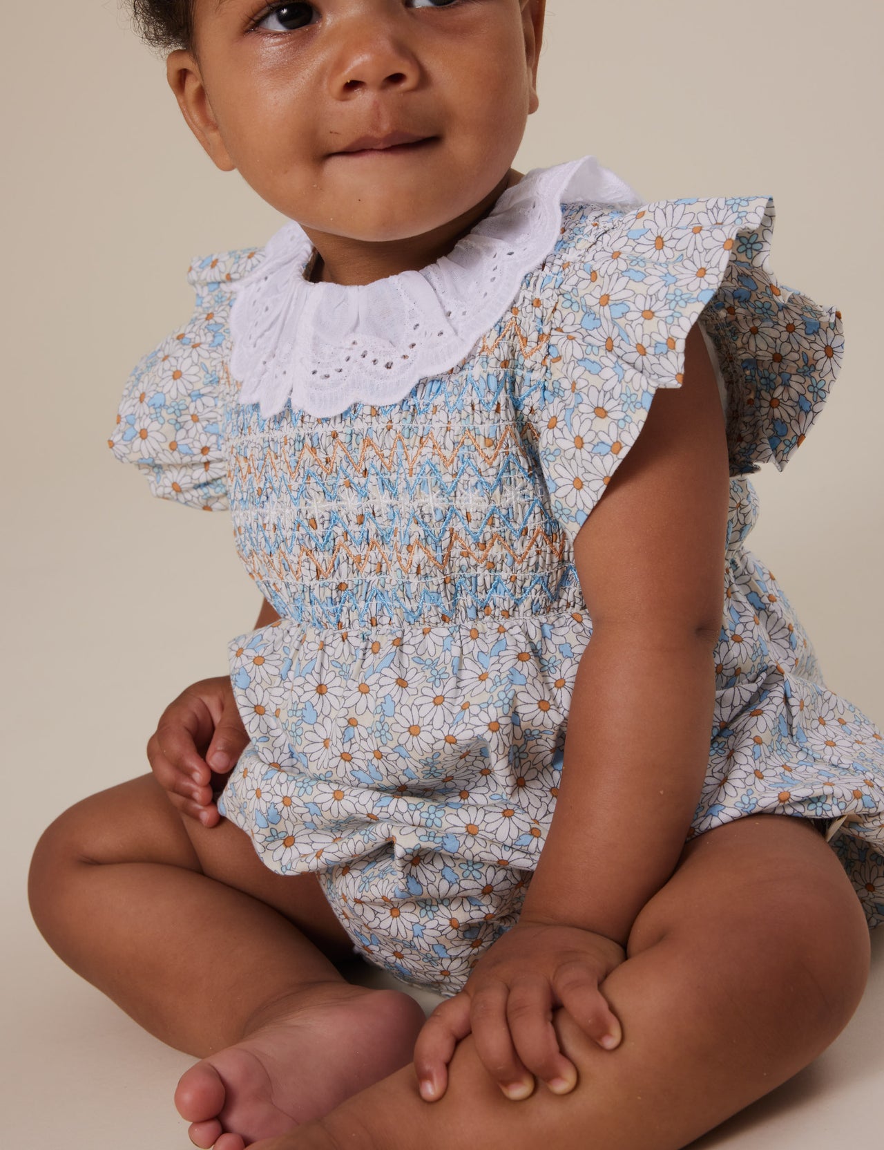 The Goldie + Ace Sunny Floral Lani Smocked Romper in Sunny Floral pictured on a grey background. The material is cotton. It is a babywear made for babies, toddlers and kids.