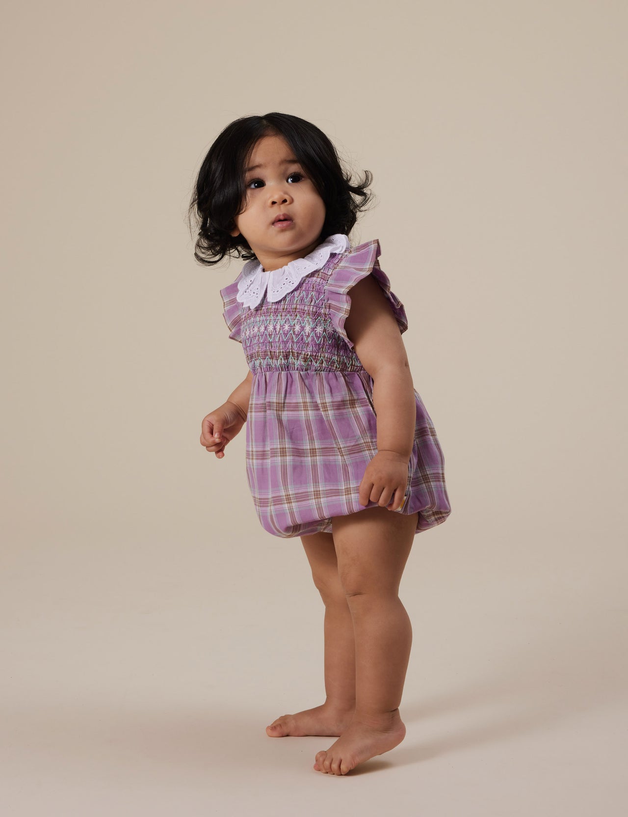 The Goldie + Ace Mauve Check Lani Smocked Romper in Mauve Check pictured on a grey background. The material is cotton. It is a babywear made for babies, toddlers and kids.