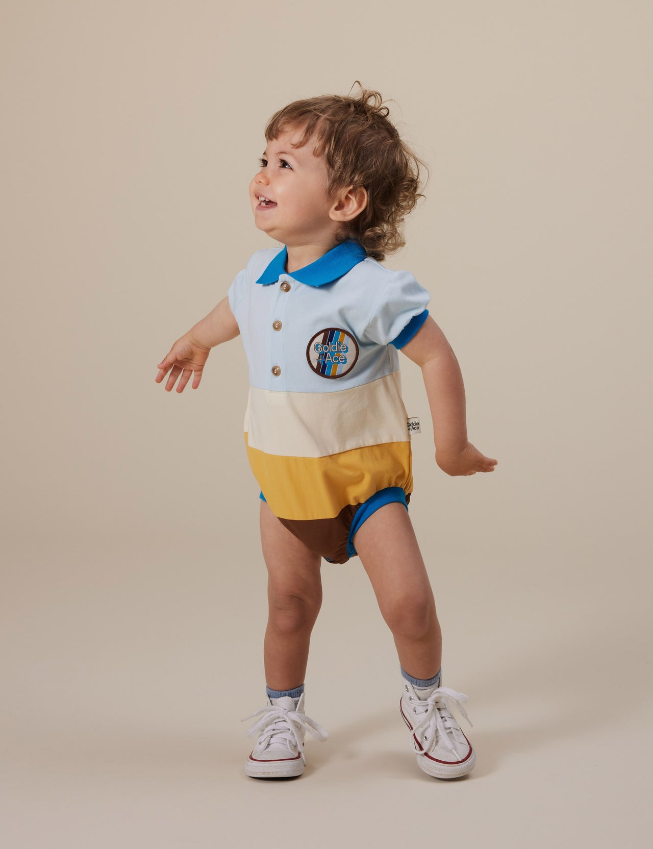 The Goldie + Ace Luca Polo Romper in Blue/Golden/Brown pictured on a grey background. The material is cotton jersey. It is a romper made for babies, toddlers and kids.