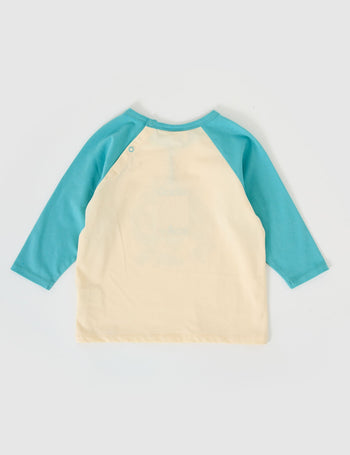 The Goldie + Ace Team Goldie Raglan Long Sleeve Top in Vanilla/Dusty Blue pictured on a grey background. The material is cotton jersey. It is a top made for babies, toddlers and kids.