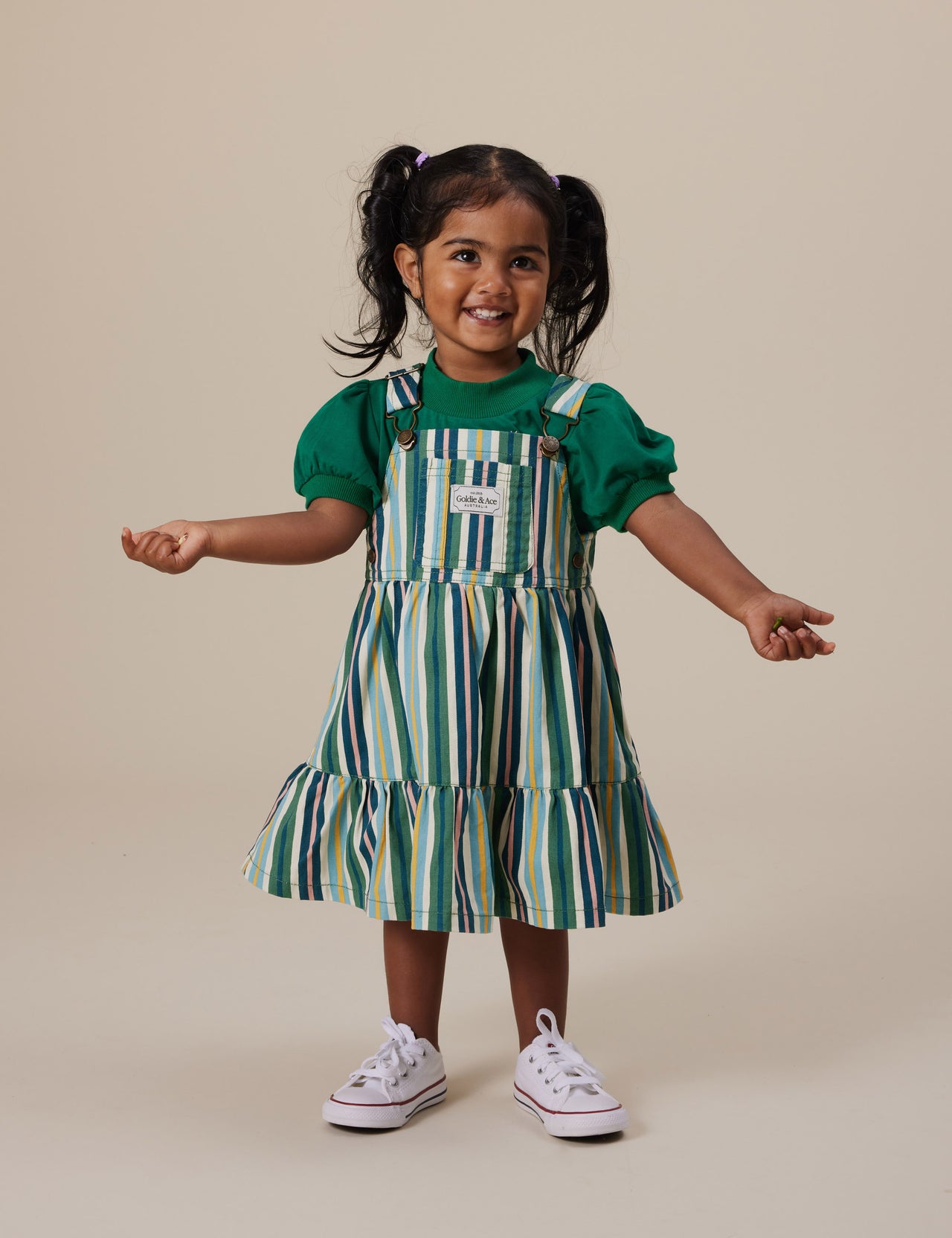 The Goldie + Ace Tully Tiered Pinafore Dress in Evergreen Stripe pictured on a grey background. The material is cotton twill. It is a dress made for babies, toddlers and kids.