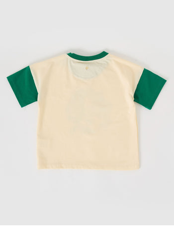 The Goldie + Ace Team Goldie T-Shirt in Vanilla/Bottle Green pictured on a grey background. The material is cotton jersey. It is a top made for babies, toddlers and kids.