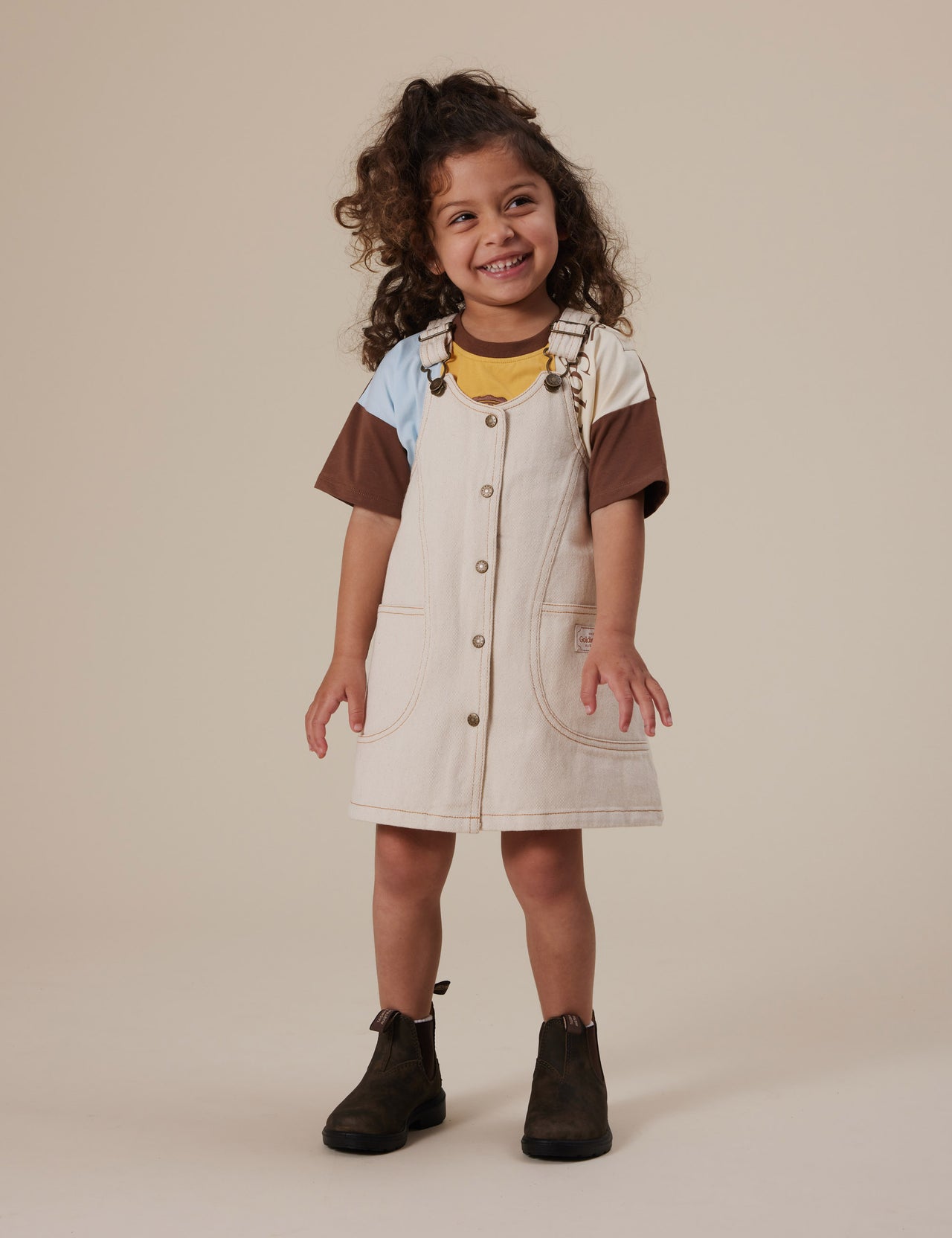 The Goldie + Ace Penelope Pinafore Pocket Dress in Oat pictured on a grey background. The material is cotton twill. It is a dress made for babies, toddlers and kids.