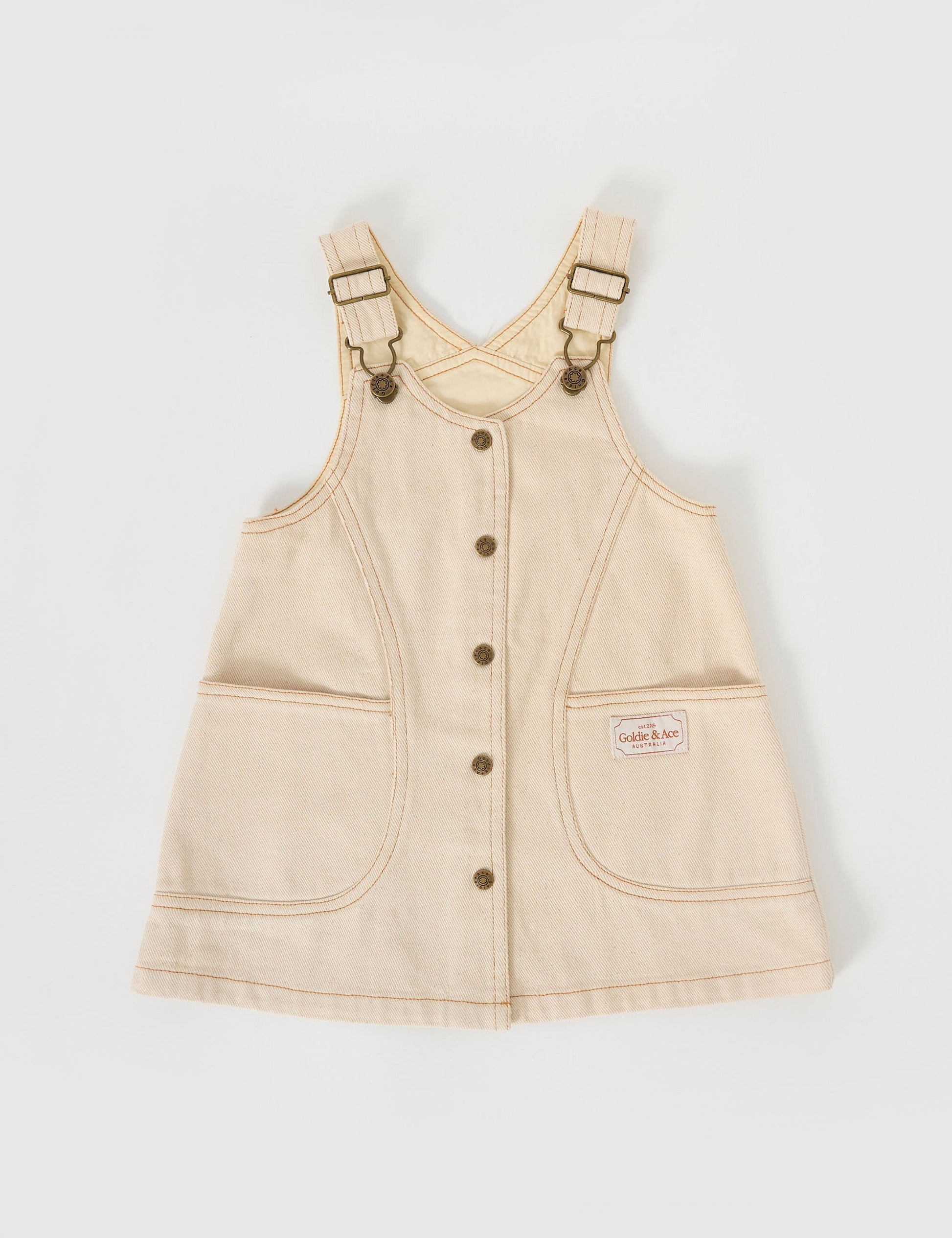 The Goldie + Ace Penelope Pinafore Pocket Dress in Oat pictured on a grey background. The material is cotton twill. It is a dress made for babies, toddlers and kids.