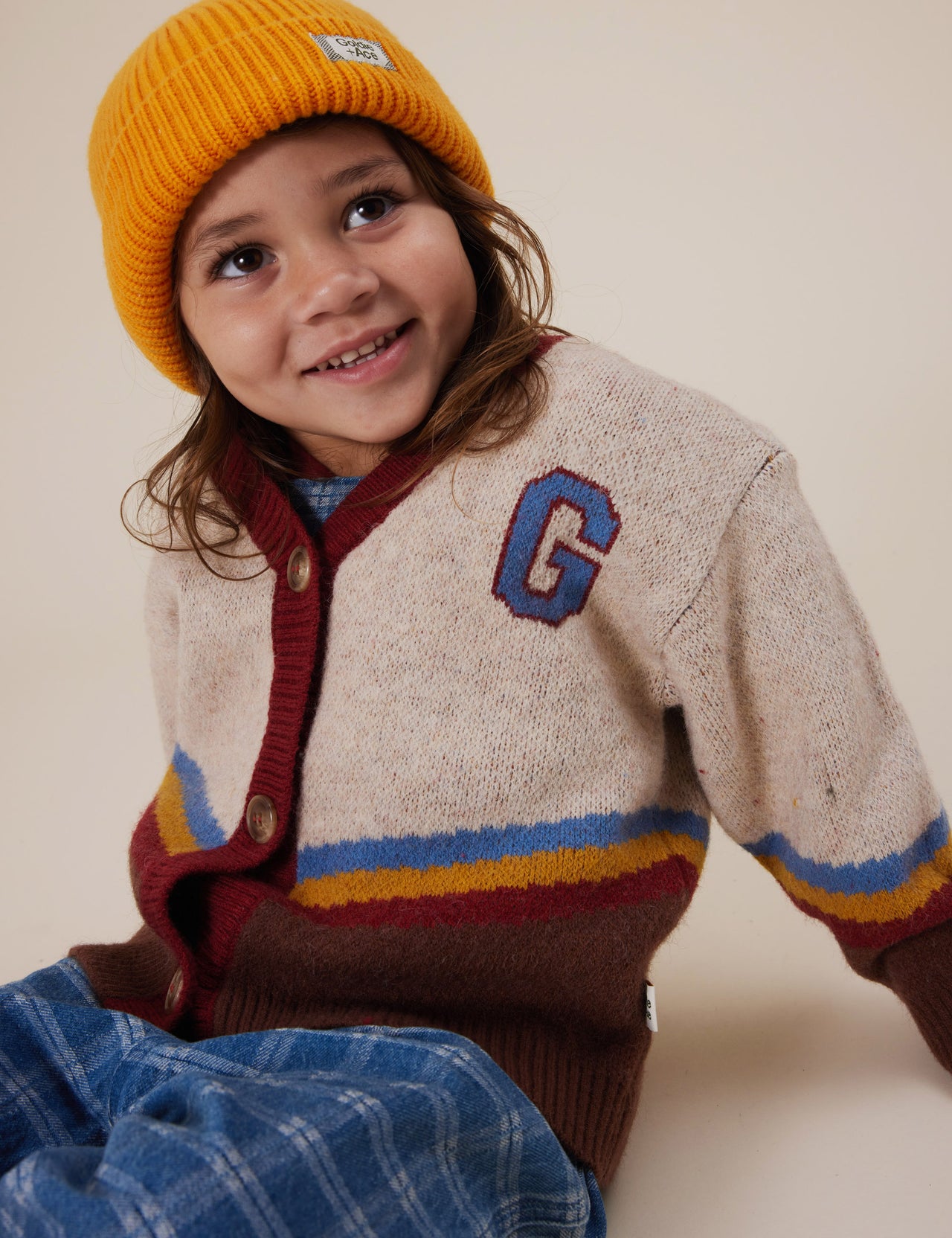 The Goldie + Ace Varsity Knit Cardigan in Vanilla/Brown pictured on a grey background. The material is super soft wool blend. It is a knit made for babies, toddlers and kids.