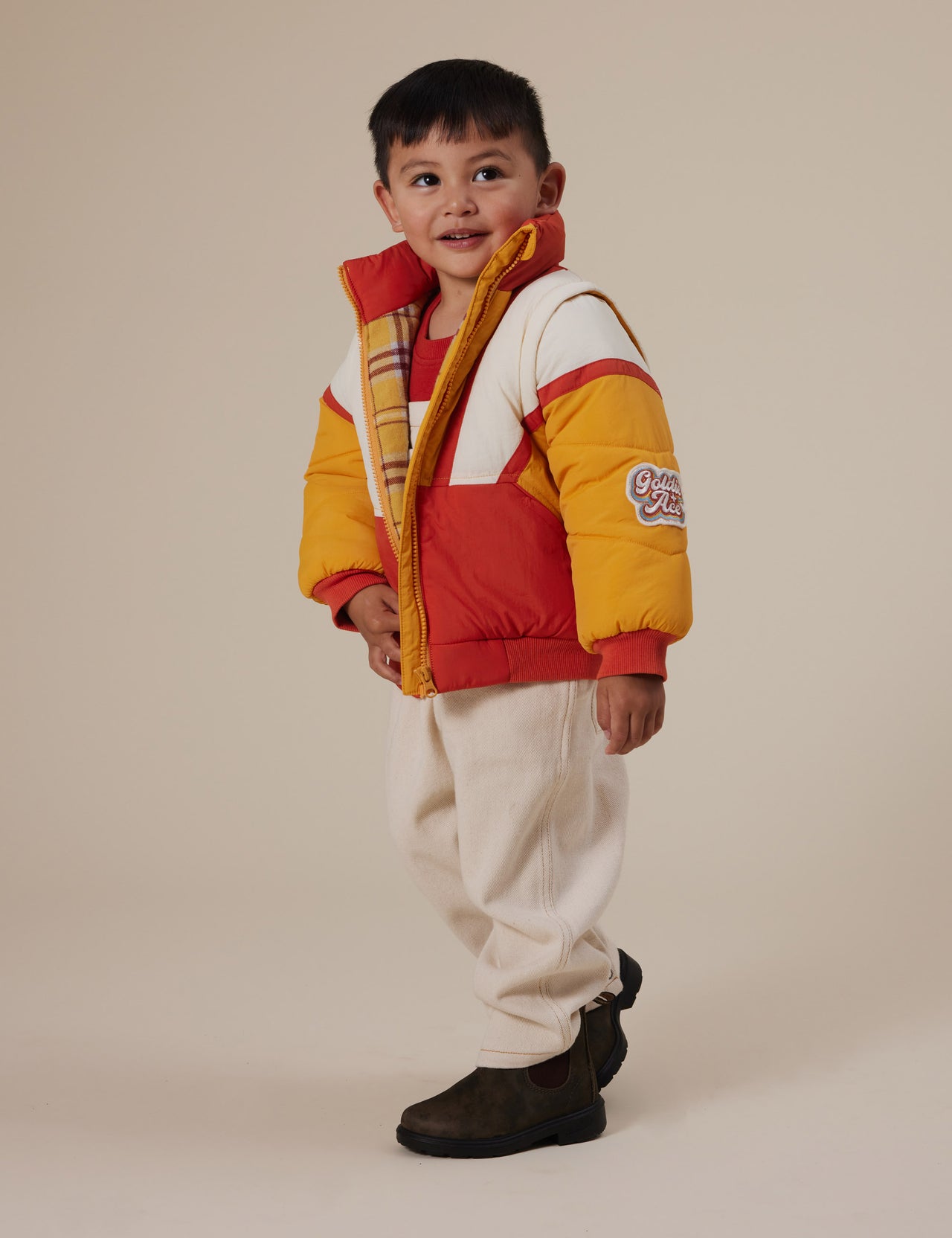 The Goldie + Ace Andy Vest/Jacket in Golden Red pictured on a grey background. The material is poly with poly fill. It is a jacket made for babies, toddlers and kids.