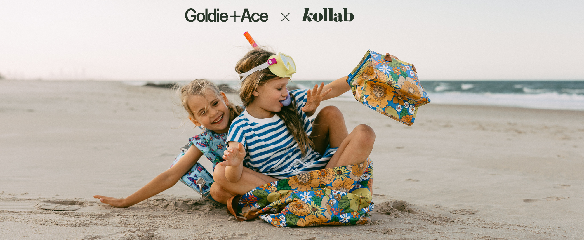 Two girls pretending to sit in a boat on the beach, wearing a striped dress and scuba goggles, and featuring a range of lunch and tote bags made by KOLLAB in collaboration with Goldie and Ace.