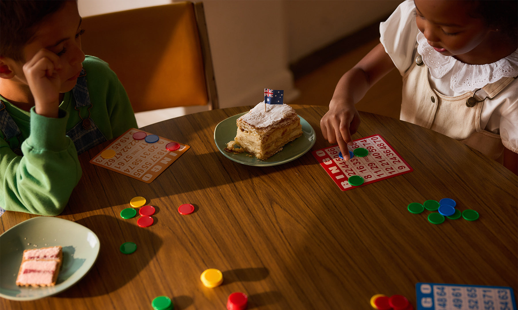 Vanilla slice with an Australia flag on a plate, next to two children playing bingo.